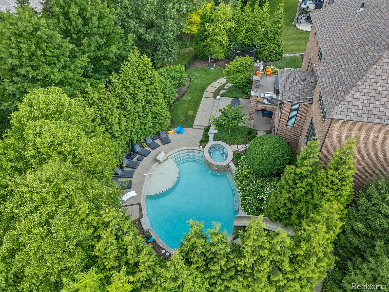 This aerial shot showcases a luxurious backyard featuring a circular pool with an attached spa, surrounded by lush greenery and mature trees. Several lounge chairs are arranged near the pool, and a patio area with outdoor furniture is visible adjacent to the brick house. The overall impression is one of a private and serene outdoor oasis.