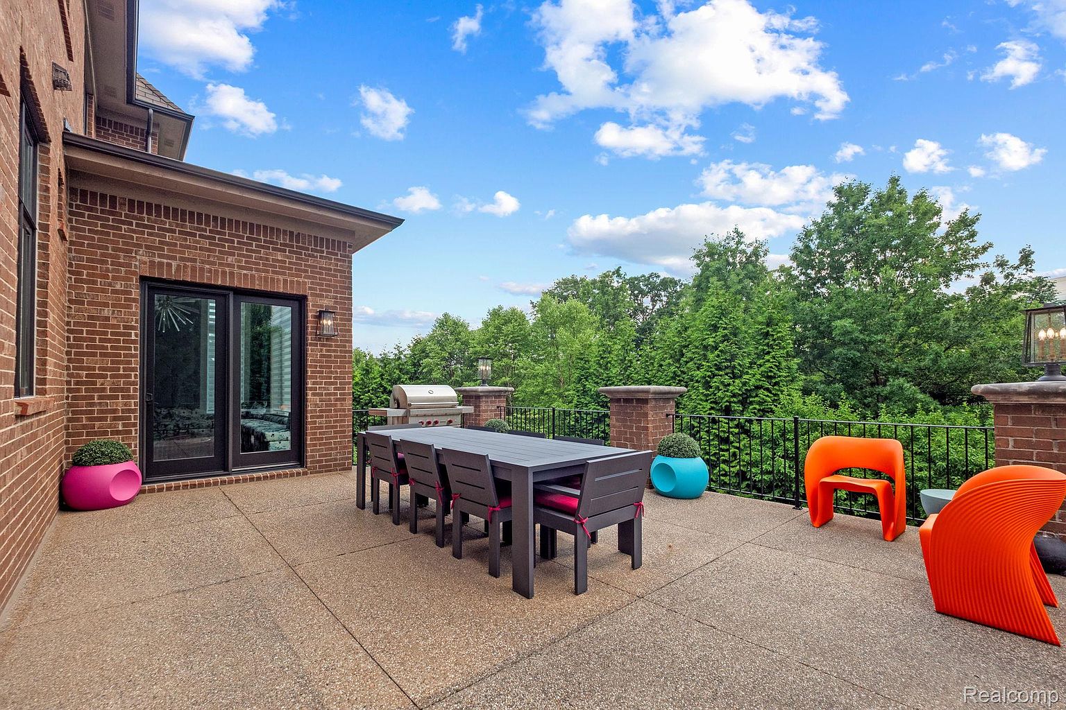This image showcases a spacious outdoor patio area, featuring a large dining table with chairs, modern orange seating, and colorful planters. The patio is constructed with a textured concrete surface and is bordered by a black metal railing, offering views of lush greenery. The area is adjacent to a brick building with glass doors, creating a seamless indoor-outdoor living experience.