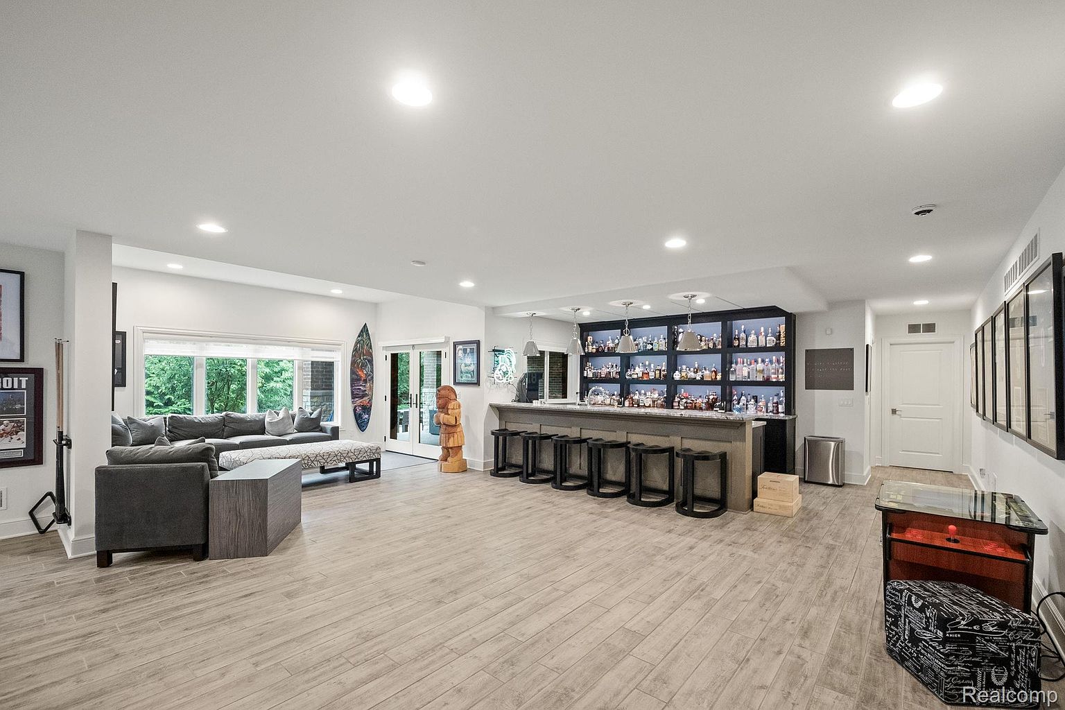 This is a spacious basement featuring a bar area with a well-stocked display, seating, and modern lighting. The room also includes a comfortable seating area with a sofa and ottoman, and the flooring is light-colored wood-look tile. The overall impression is a stylish and inviting entertainment space.