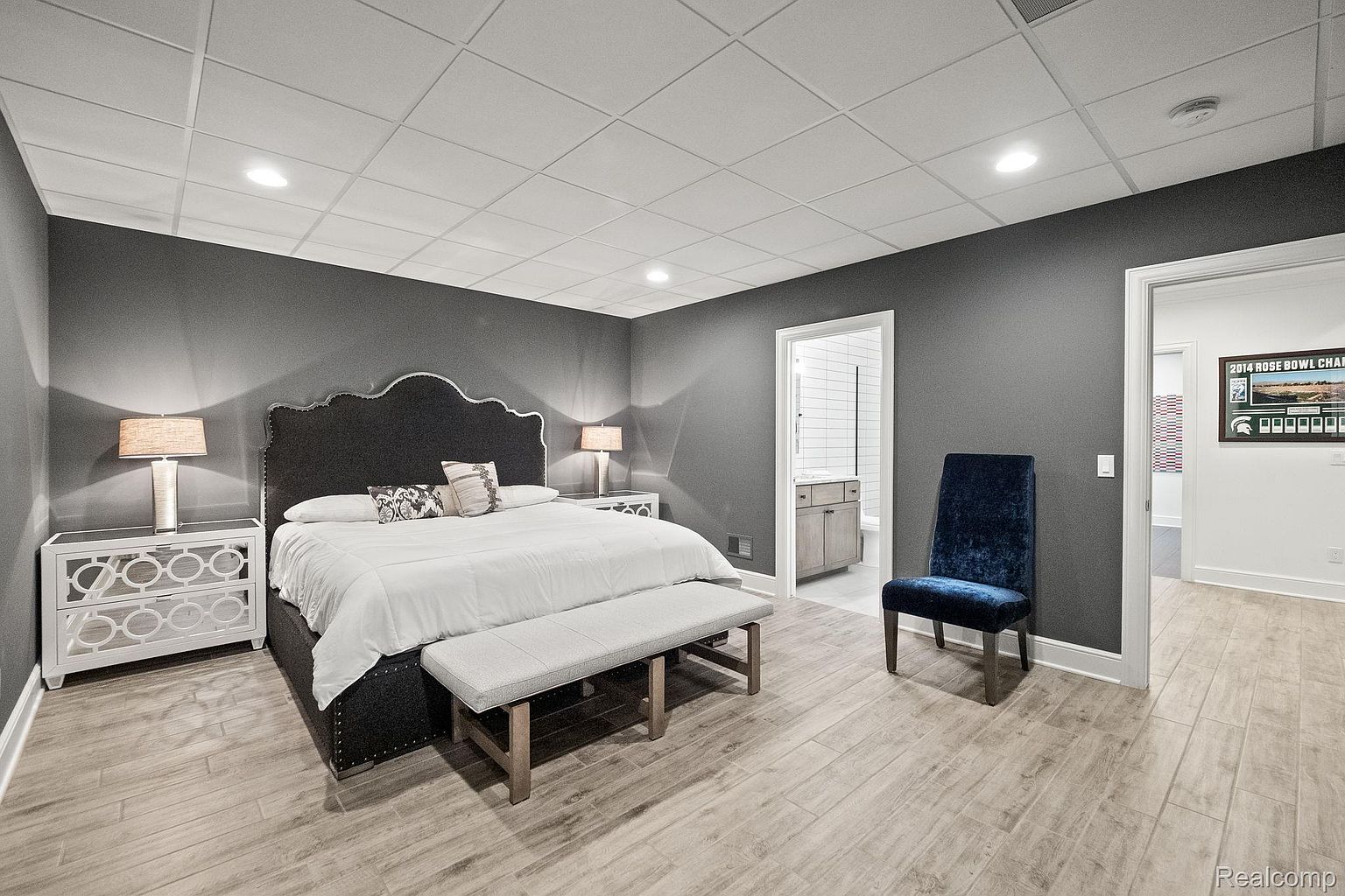 This is a primary bedroom featuring a dark gray color scheme with a large upholstered headboard and a white comforter. The room includes two white nightstands with lamps, a gray upholstered bench at the foot of the bed, and a blue velvet chair. The flooring is light wood-look tile, and there are doorways leading to a bathroom and another room, creating a spacious and inviting atmosphere.