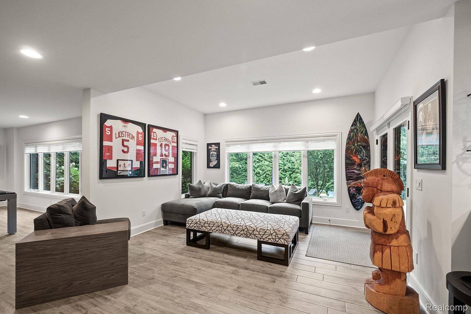 This is a well-lit living room featuring a large sectional sofa with decorative pillows, a patterned ottoman, and a wooden accent chair. Framed sports jerseys hang on the wall, adding a personalized touch. A wooden tiki statue and surfboard add a unique decorative element, while large windows provide natural light and views of the greenery outside.