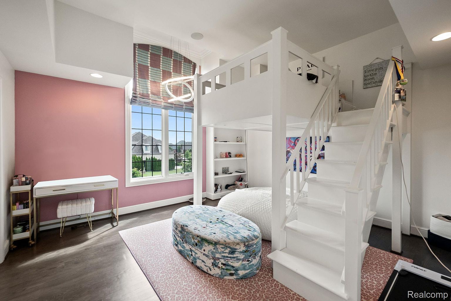 This is a bright and stylish bedroom featuring a unique loft bed with stairs, a desk area with a vanity, and a patterned rug. The room is painted in a combination of pink and white, creating a cheerful and inviting atmosphere. The large window provides ample natural light and a view of the outdoors.