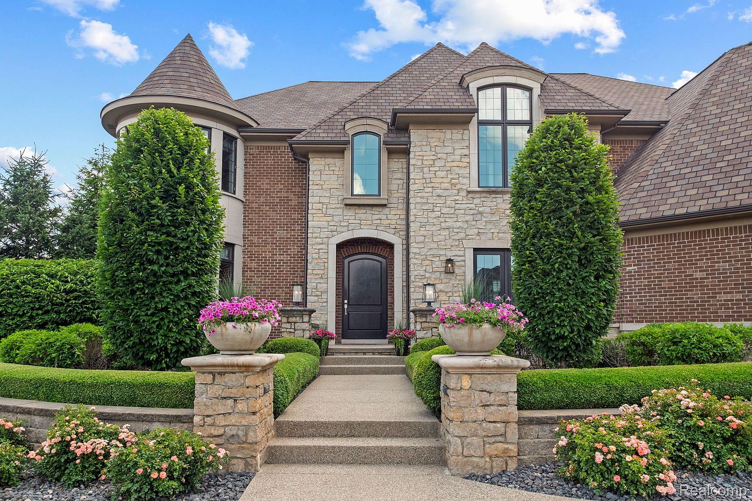 This is a front view of a luxurious home showcasing a grand entryway. The facade combines stone and brick elements, complemented by manicured greenery and flower arrangements. A paved walkway leads to a dark, arched front door, creating an inviting and upscale impression.