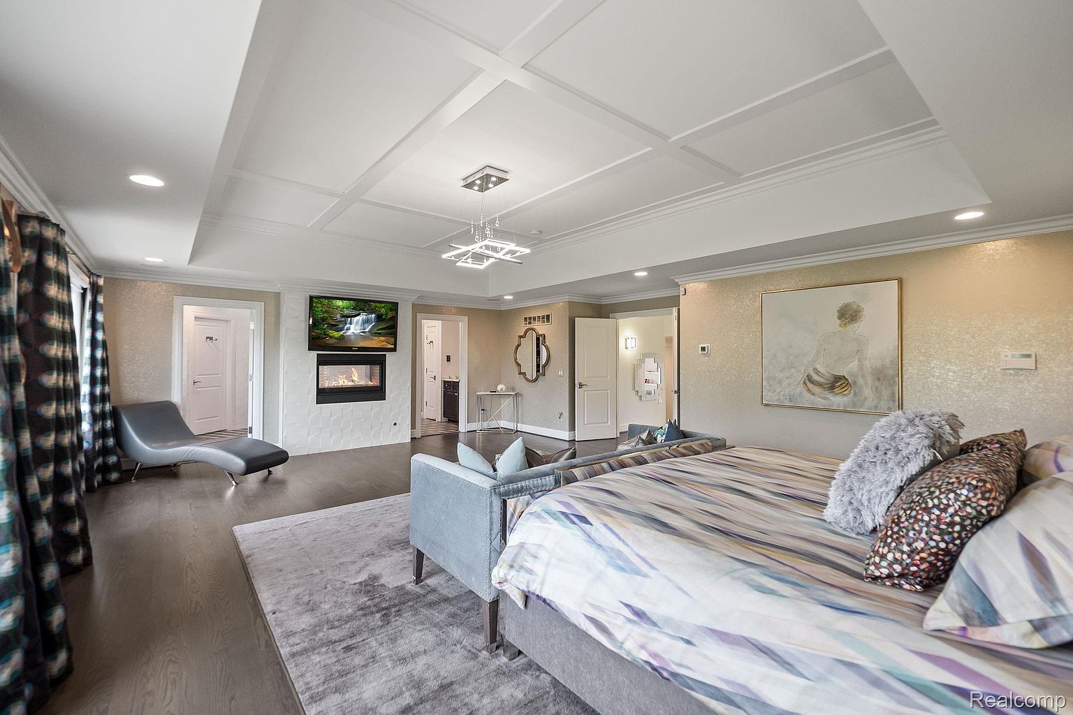 This is a luxurious primary bedroom featuring a coffered ceiling with a modern chandelier, a fireplace with a TV mounted above, and a large bed with decorative pillows. The room is decorated in neutral tones with a statement wall art piece, creating a serene and elegant atmosphere. The perspective is from the foot of the bed, showcasing the room's depth and features.