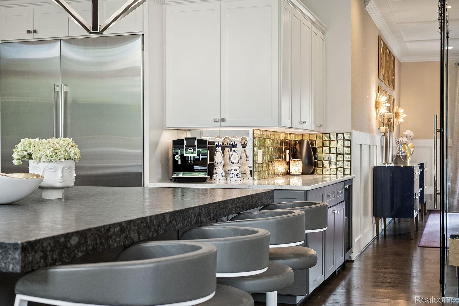 This is a well-lit kitchen featuring a dark countertop with barstool seating, white upper cabinets, and gray lower cabinets. The backsplash is a reflective tile, and stainless steel appliances are visible. The kitchen has a modern and upscale feel, with decorative items adding a touch of elegance.