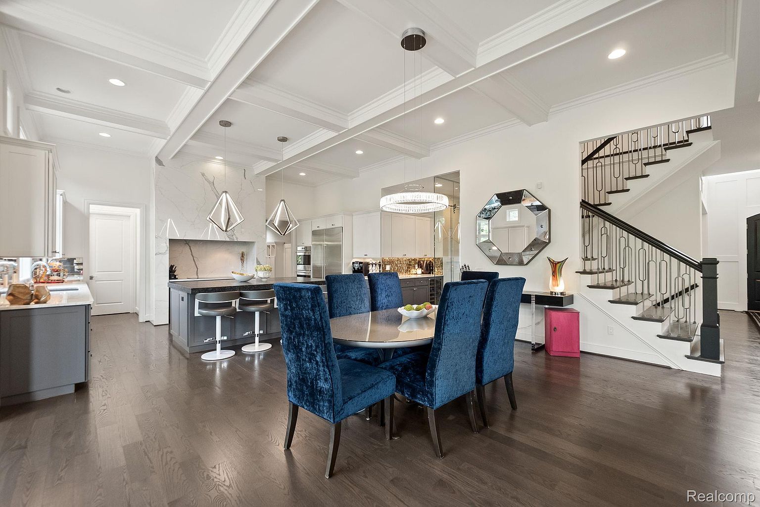 This interior shot showcases a modern dining area adjacent to a kitchen. The dining space features a round table surrounded by six plush, blue velvet chairs. A striking chandelier hangs above the table, and a unique octagonal mirror adorns the wall, reflecting light and adding visual interest. The dark hardwood floors provide a rich contrast to the white walls and ceiling, creating a sophisticated and inviting atmosphere.