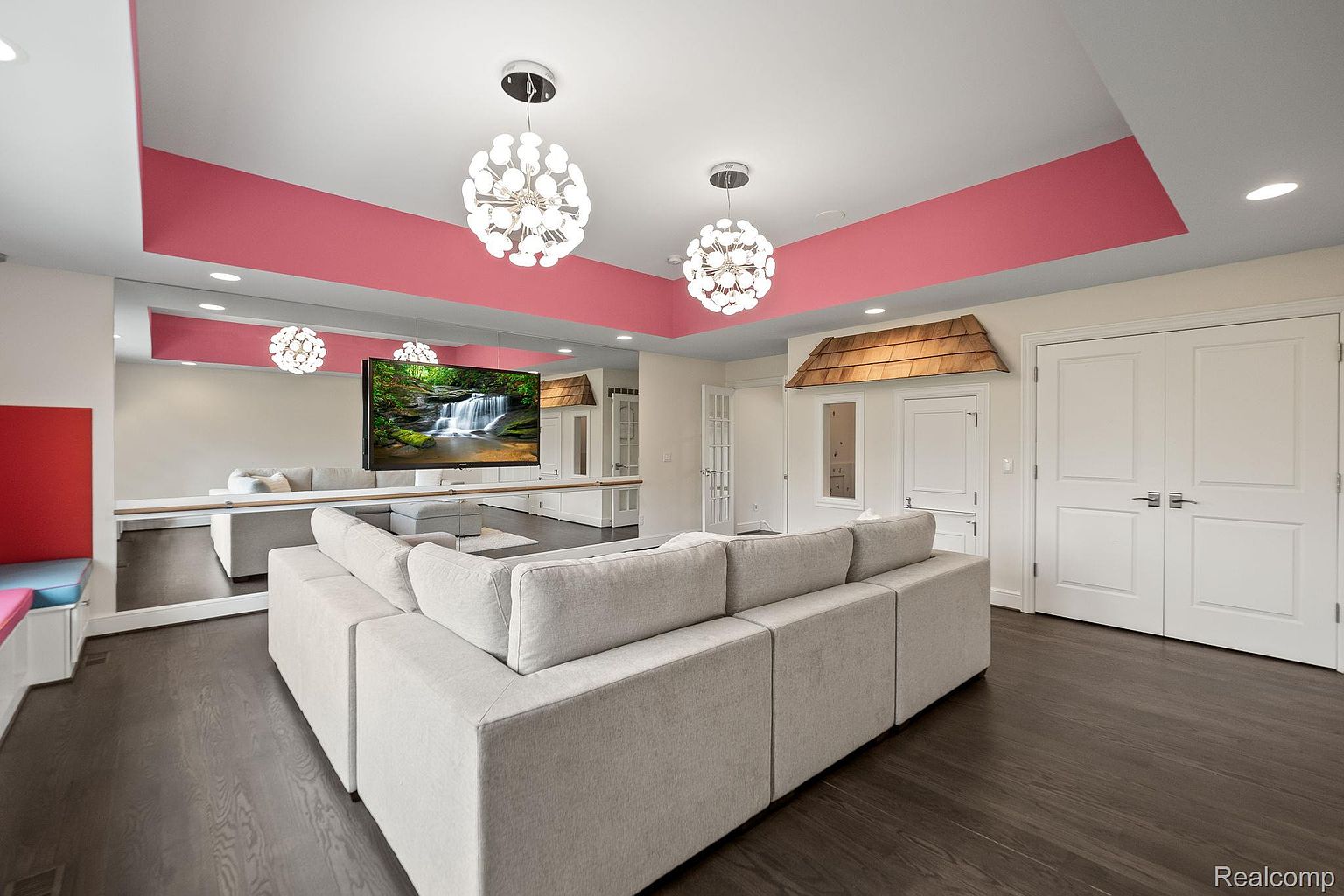 This is an interior shot of a modern living room featuring a large sectional sofa in a neutral tone, dark hardwood floors, and unique lighting fixtures. A large mirror spans the length of one wall, reflecting the sofa and a mounted television displaying a waterfall scene. The room also includes a playful architectural detail resembling a small house facade with a door and window, adding a whimsical touch.