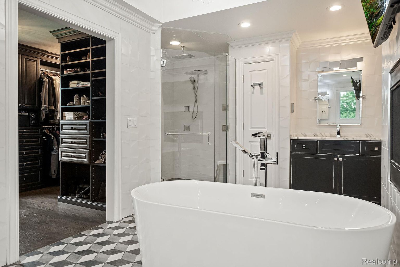 This is a luxurious primary bathroom featuring a freestanding white bathtub as a focal point. The room includes a glass-enclosed shower, a dark vanity with a mirror, and a walk-in closet visible through an open doorway. The flooring is a geometric pattern, and the walls are tiled, creating a clean and modern aesthetic.