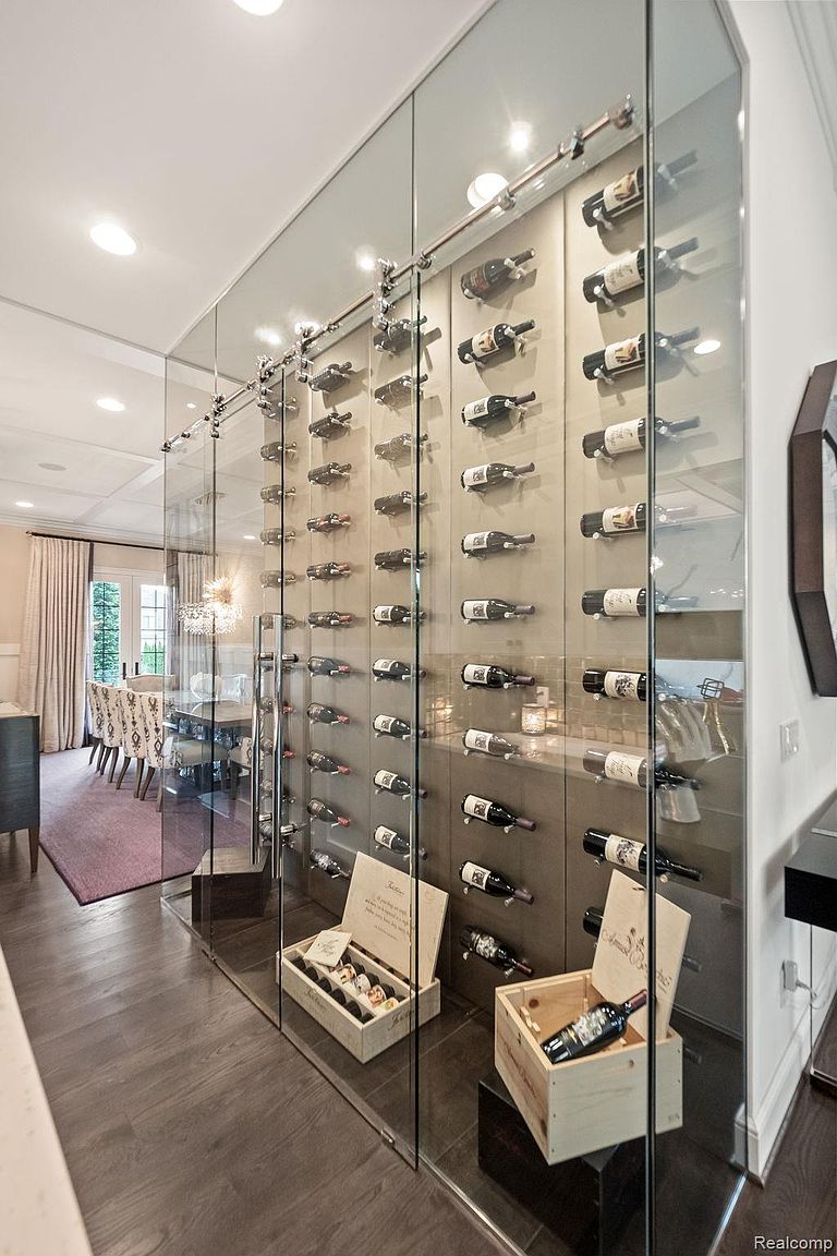 This image showcases a modern wine cellar with a glass enclosure, displaying numerous wine bottles horizontally on a wall-mounted system. The cellar features wooden wine crates and a dark wood floor, creating a sophisticated and temperature-controlled environment for wine storage. The dining room is visible through the glass, suggesting a seamless transition between entertaining spaces.