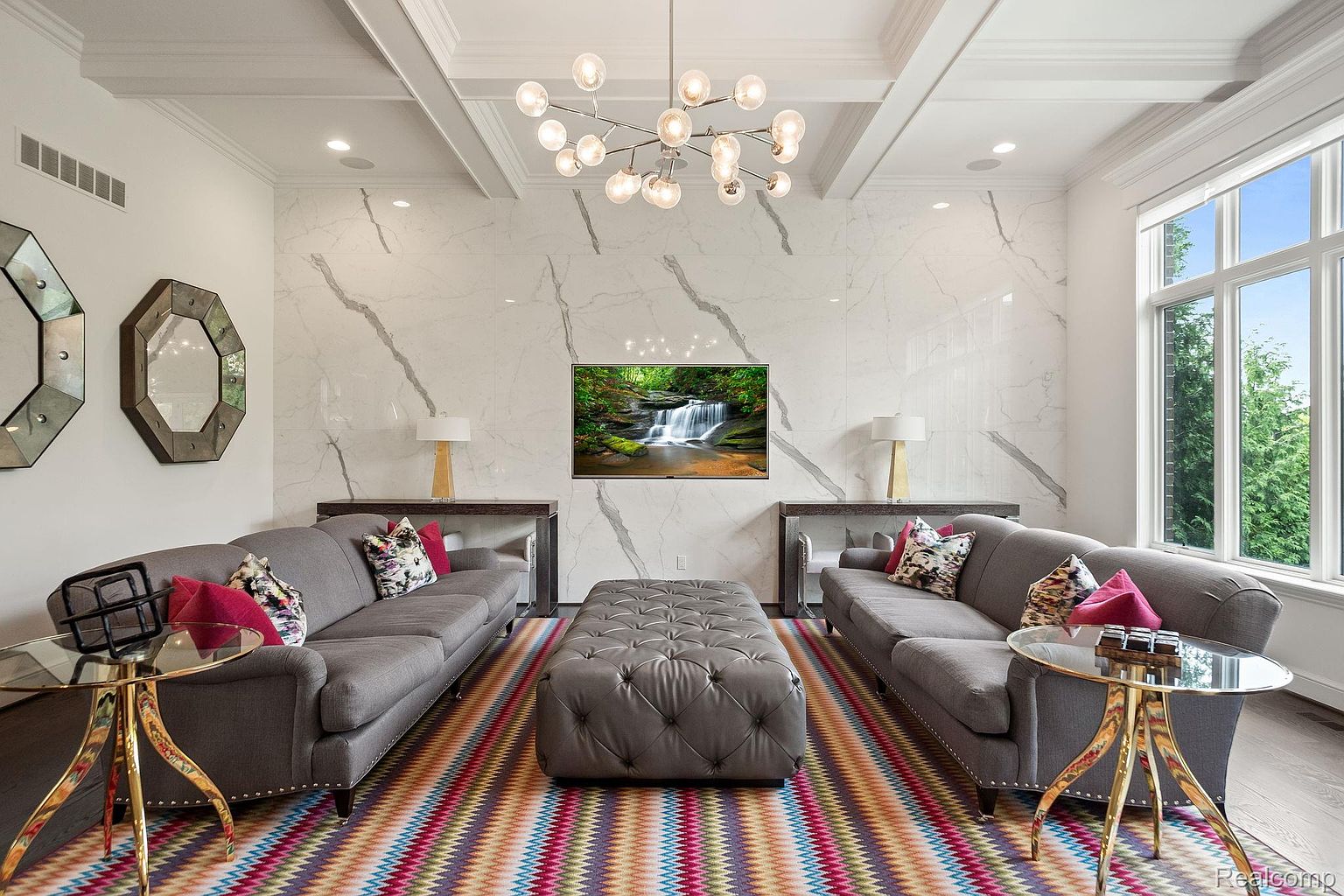 This is an interior shot of a luxurious living room featuring two gray sofas facing each other, separated by a tufted ottoman. A colorful chevron patterned rug adds vibrancy to the space, while a modern chandelier hangs from the coffered ceiling. The room is decorated with marble-like wall panels and includes side tables with glass tops and gold legs, creating an elegant and inviting atmosphere.
