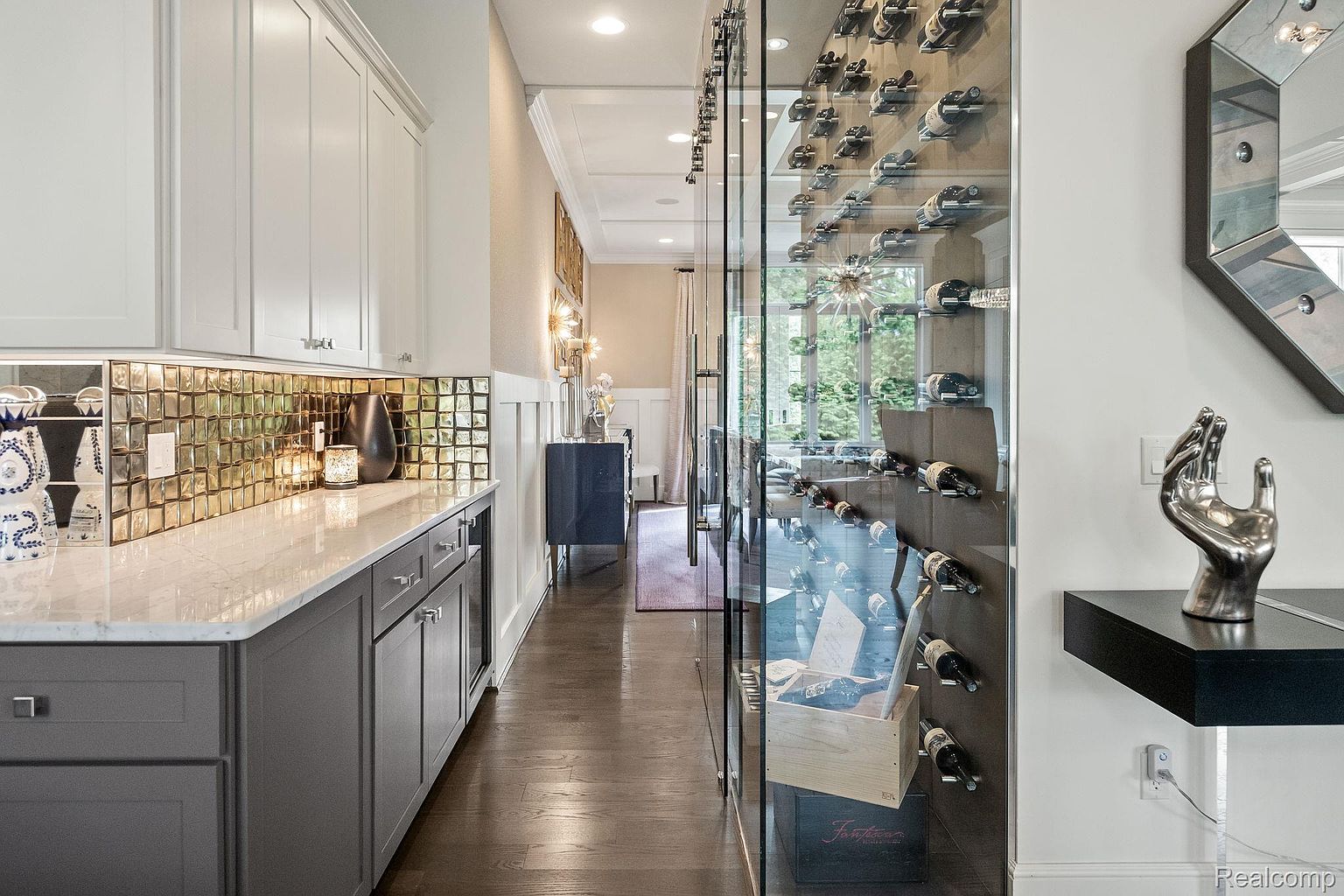 This image showcases a sophisticated wine cellar, featuring a glass-enclosed wine rack filled with bottles. The adjacent area includes elegant gray cabinetry with a marble countertop and a mirrored tile backsplash. A decorative silver hand sculpture sits on a black floating shelf, adding a touch of modern art.