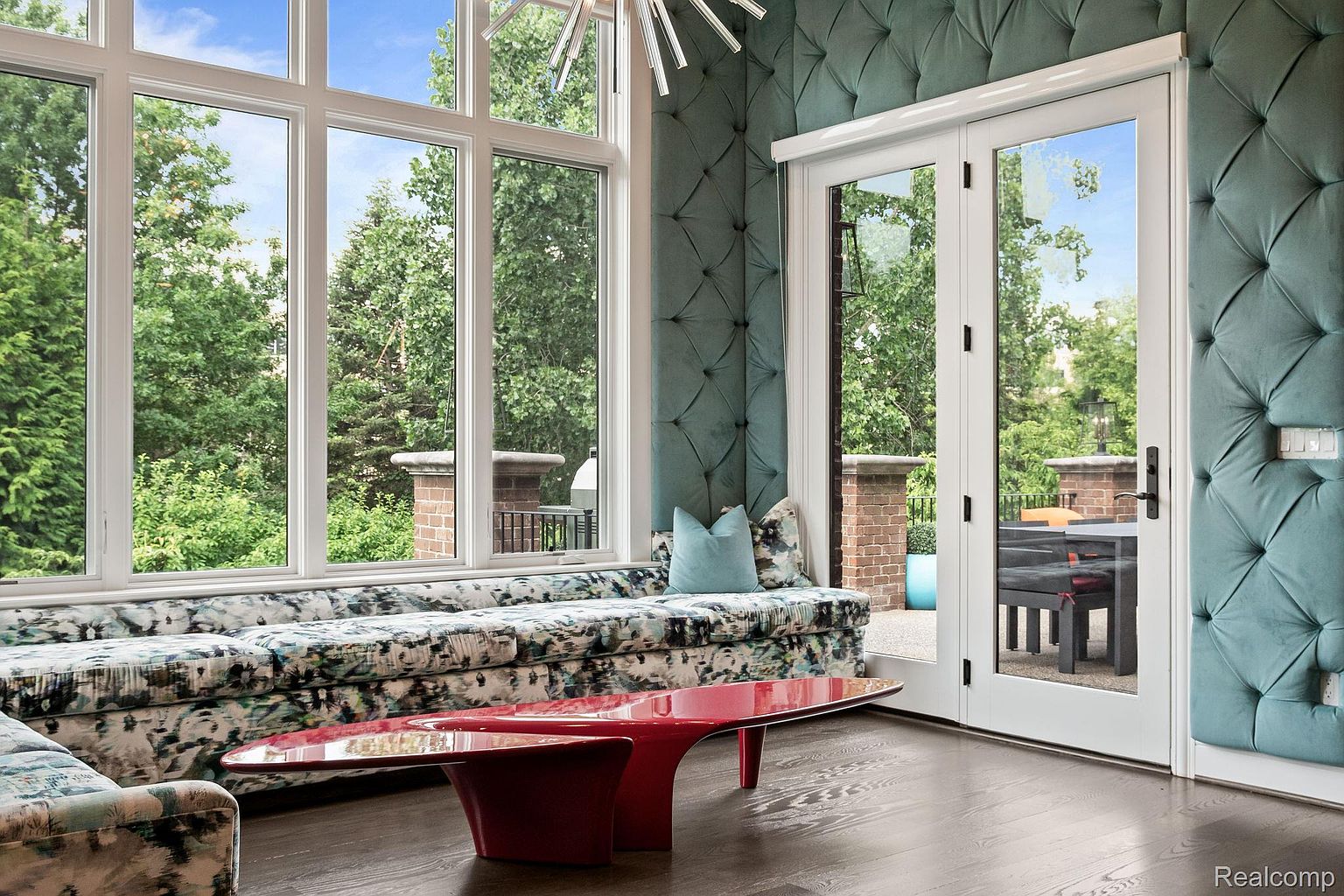 This interior shot showcases a luxurious living room with a large window offering a view of lush greenery. The room features a unique, patterned sofa, a modern red coffee table, and tufted teal wall panels, creating a sophisticated and stylish atmosphere. The hardwood floors add warmth and elegance to the space.