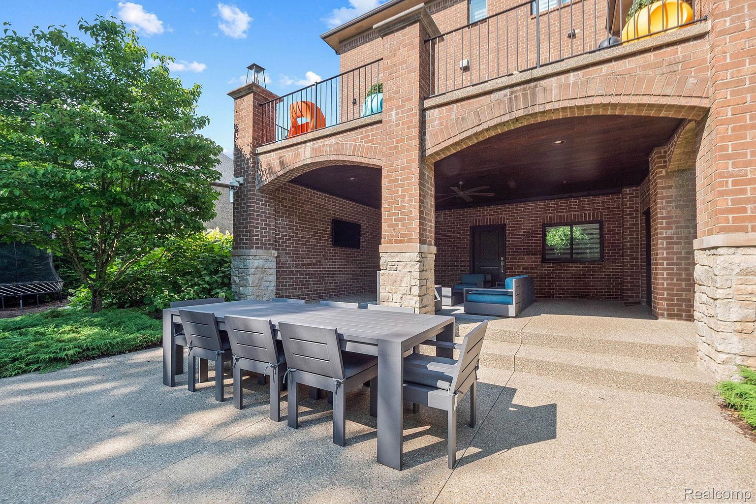This image showcases a luxurious outdoor patio area, featuring a large dining table with seating for eight, set against the backdrop of a brick-clad two-story structure. The patio is paved with textured concrete, and the upper level includes a balcony with wrought iron railings. The space is designed for outdoor entertaining and relaxation, offering a seamless transition between indoor and outdoor living.