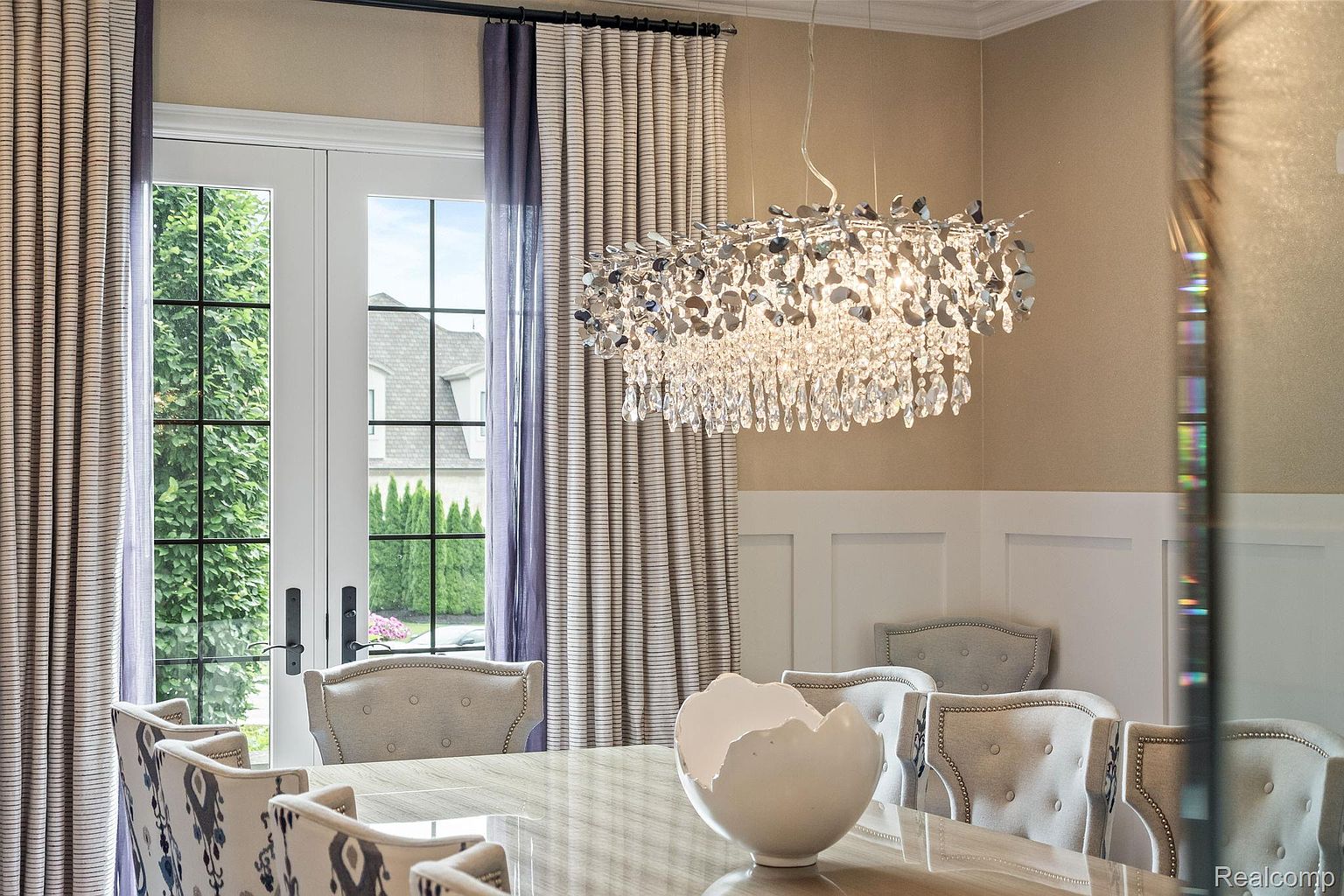 This is an interior shot of a dining room, showcasing a modern chandelier hanging above a glossy dining table with a decorative white bowl centerpiece. The room features elegant chairs with button detailing, complemented by striped and sheer curtains framing a window view of the outdoors. The wainscoting on the walls adds a touch of traditional charm to the contemporary design.
