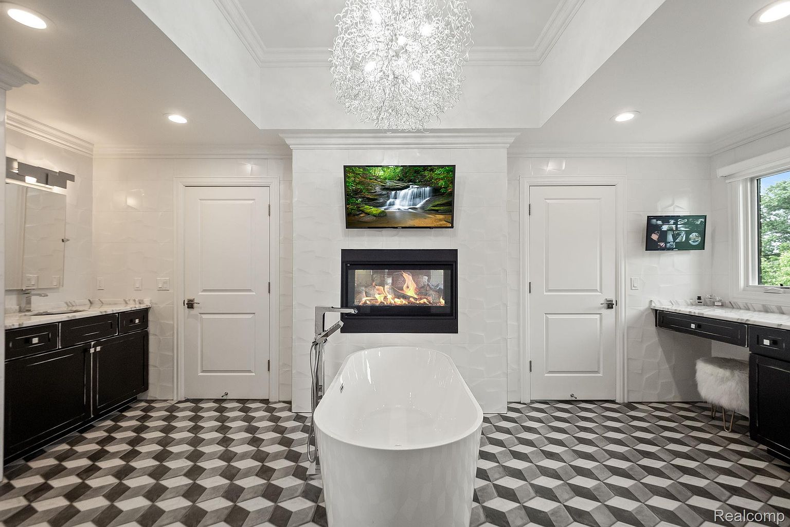 This is a luxurious primary bathroom featuring a freestanding white bathtub as a focal point, complemented by a modern fireplace and a wall-mounted television. The room is decorated with a geometric-patterned floor, white textured walls, and dark cabinetry, creating a sophisticated and relaxing atmosphere. A chandelier hangs from the ceiling, adding a touch of elegance.