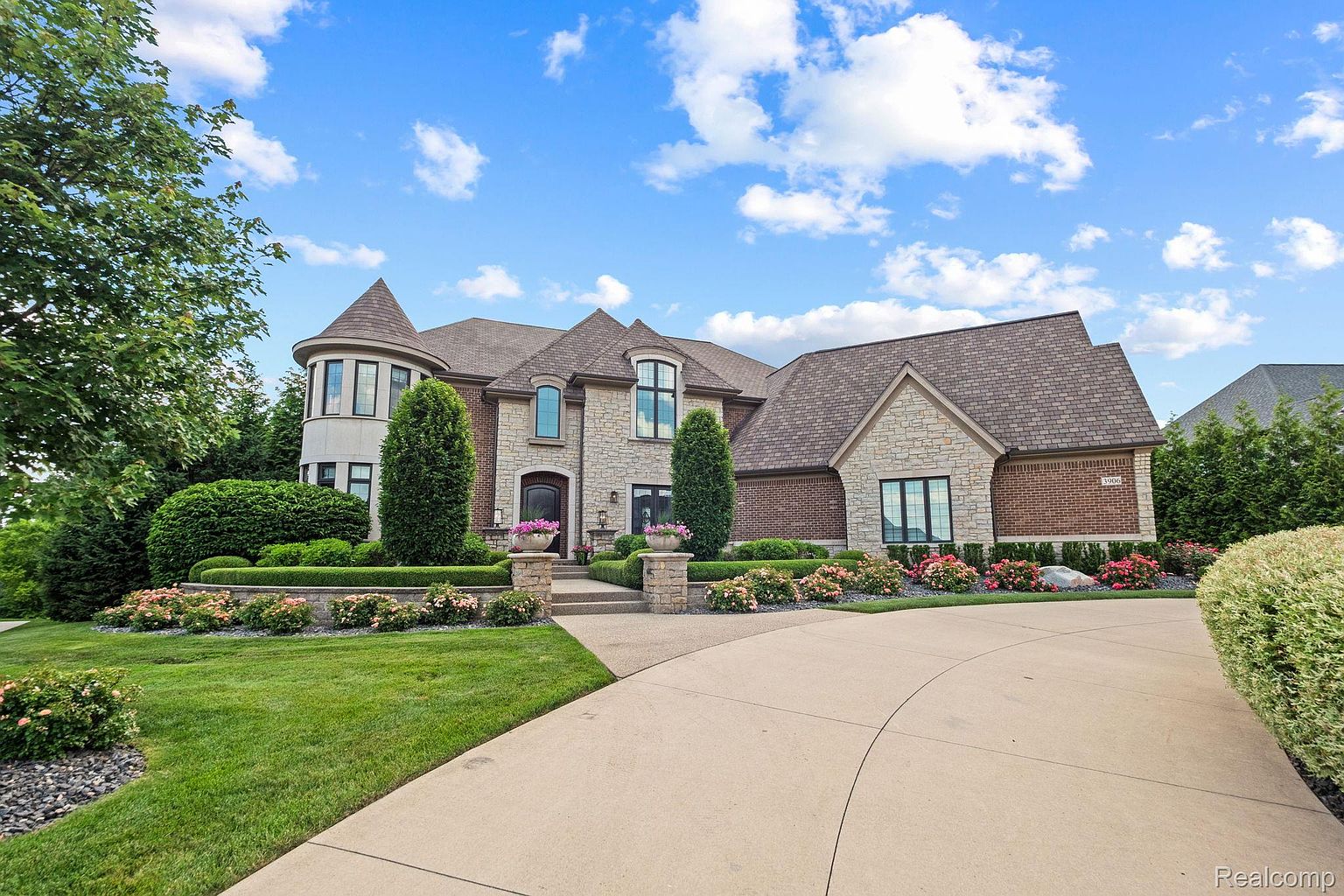 This is a front view of a large, luxurious home with a well-manicured lawn and landscaping. The house features a combination of stone and brick exterior, a turret-style architectural element, and a winding driveway leading to the entrance. The overall impression is one of elegance and curb appeal.