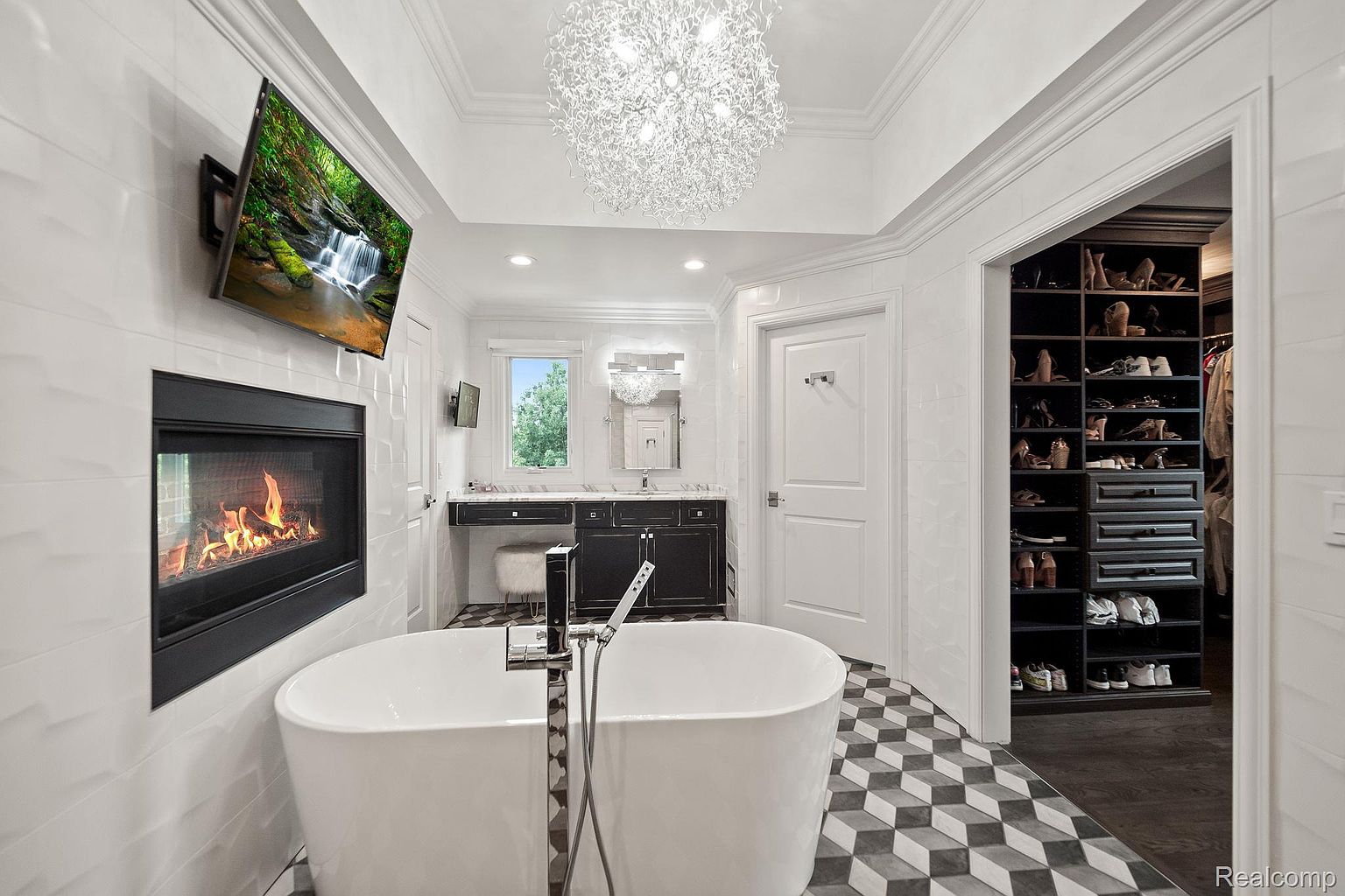 This is a luxurious primary bathroom featuring a freestanding white bathtub with a modern faucet, a fireplace with a TV above it, and a dazzling chandelier. The black and white geometric tile flooring adds a touch of sophistication, while the adjacent walk-in closet provides ample storage. The overall impression is one of elegance and relaxation.
