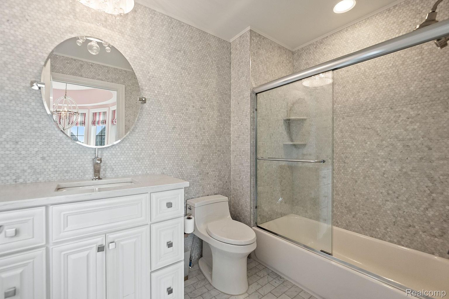This is a bathroom featuring a white vanity with a round mirror above it, a toilet, and a bathtub with a glass shower door. The walls are covered in a small, light-colored mosaic tile, creating a clean and modern look. The overall impression is a well-maintained and stylish bathroom.