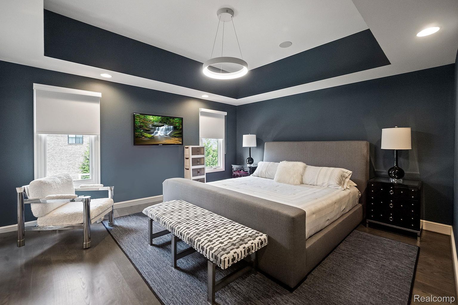 This is a primary bedroom featuring a gray upholstered bed with a bench at the foot, set on a gray rug. The walls are painted a dark blue, and there's a modern circular light fixture hanging from the ceiling. A TV is mounted on the wall, and there are windows with white blinds on either side of the bed.