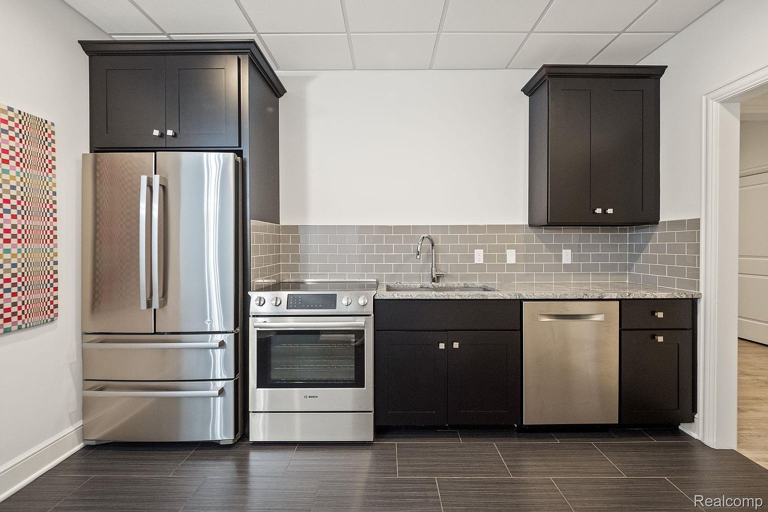 This is a well-appointed kitchen area featuring stainless steel appliances including a refrigerator, oven, and dishwasher. Dark cabinetry contrasts with the light gray backsplash and countertop, creating a modern aesthetic. The dark tiled flooring adds a touch of sophistication to the space.