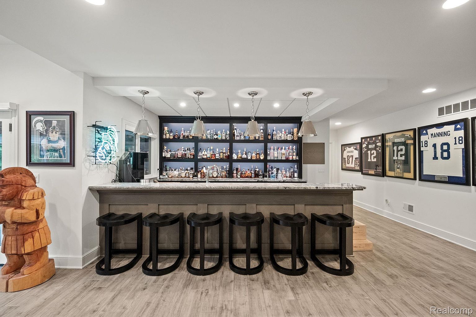 This is an interior shot of a finished basement featuring a stylish bar area. The bar has a gray countertop, dark wood cabinetry, and black bar stools. Behind the bar is a well-stocked liquor cabinet with lighting, and sports memorabilia adorns the walls, creating a relaxed and entertaining atmosphere.