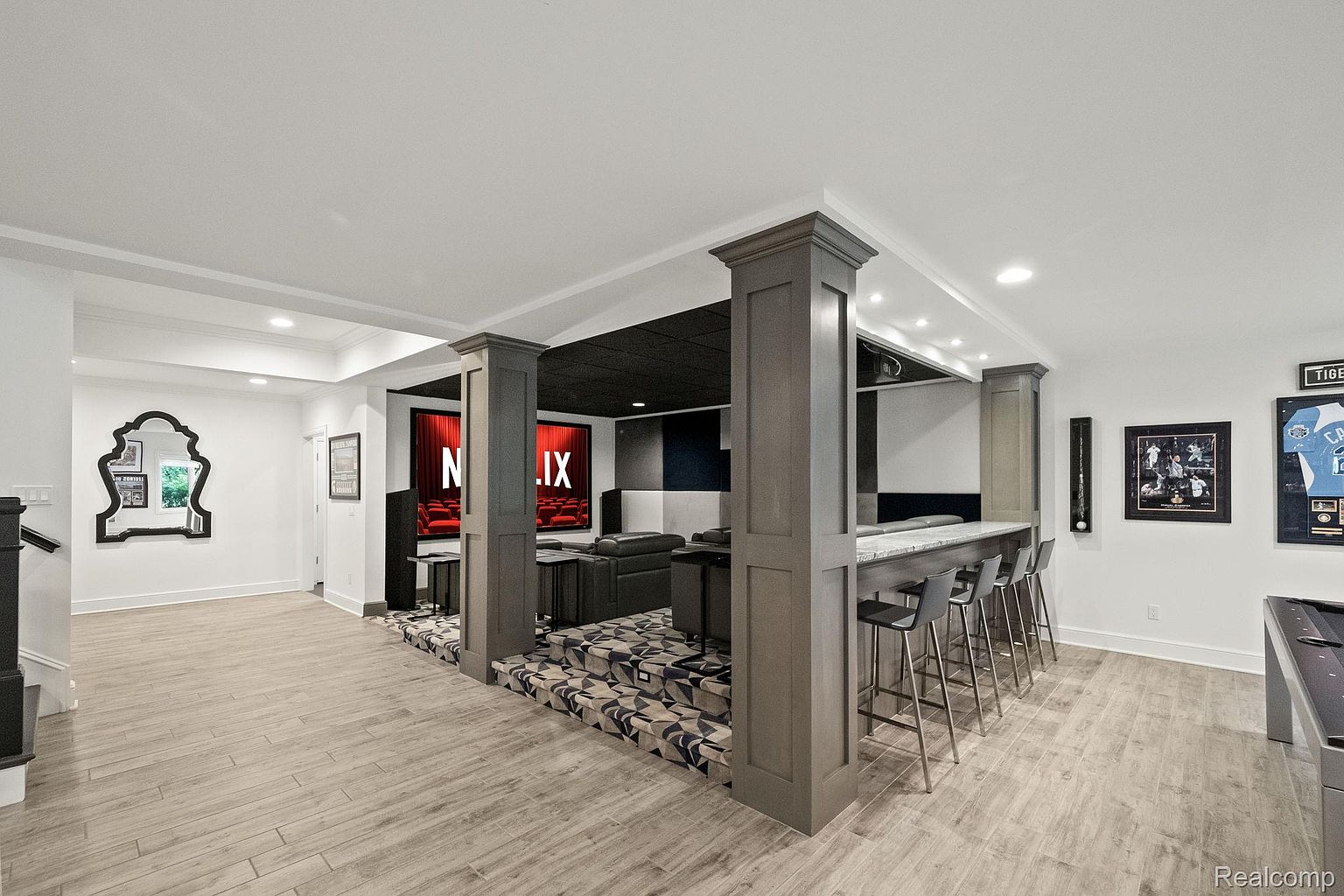 This is an interior shot of a finished basement featuring a home theater and bar area. The theater has tiered seating and a large screen, while the bar includes counter seating and custom cabinetry. The flooring is a light wood-look tile, and the overall design is modern and inviting.