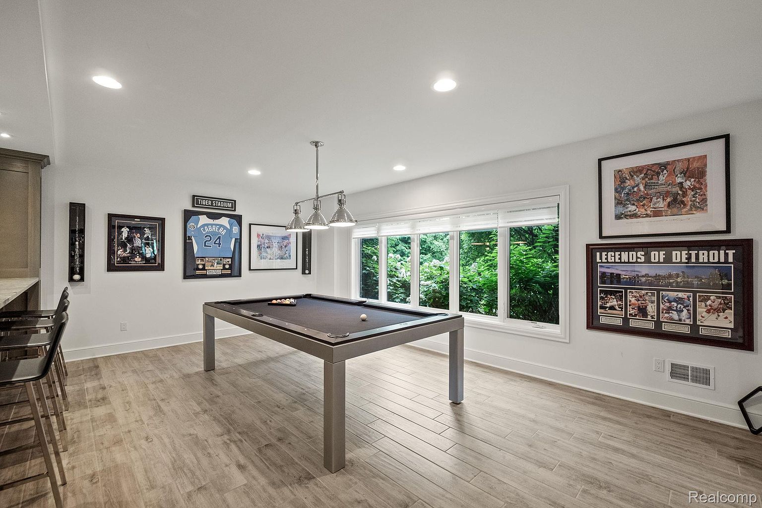 This is a well-lit basement featuring a modern pool table as the focal point. The room is decorated with sports memorabilia, including framed jerseys and artwork, adding a personalized touch. The light wood-look tile flooring and neutral wall colors create a clean and inviting atmosphere, enhanced by natural light from the window.