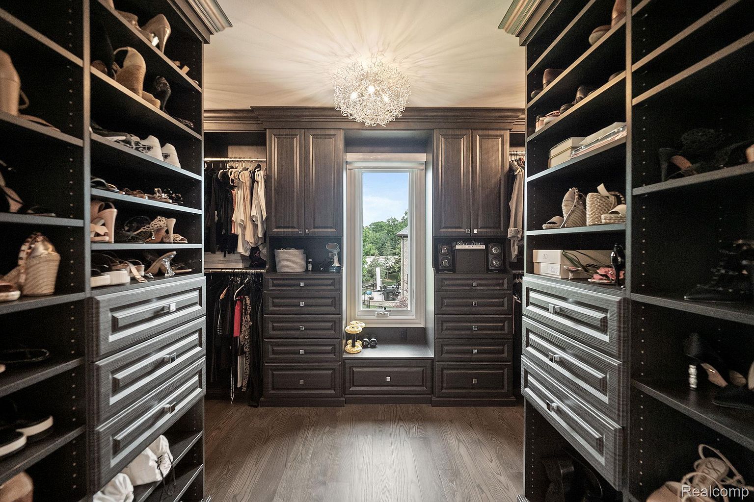 This is a well-organized walk-in closet featuring custom dark wood cabinetry and shelving. The closet is designed with ample storage for shoes, clothing, and accessories, and a window provides natural light. A decorative chandelier adds a touch of elegance to the space.