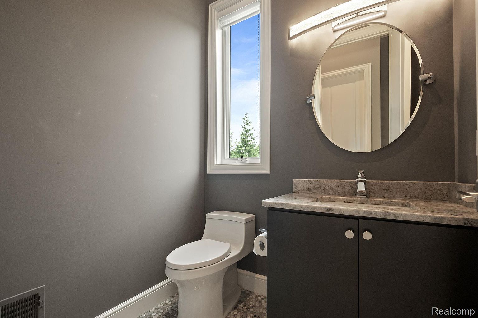 This is a well-lit guest bathroom featuring a modern design. The room has a gray color scheme, a round mirror above a dark vanity with a light countertop, and a toilet next to a window. The flooring appears to be a mosaic tile, adding a touch of elegance.