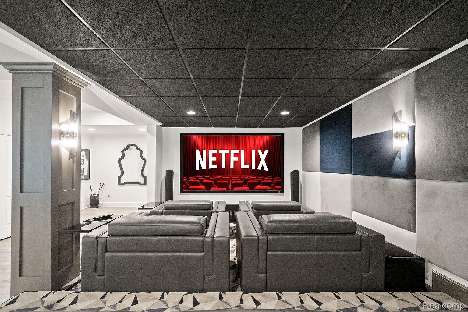 This is a well-designed home theater in a basement, featuring tiered seating with gray leather recliners, a large screen displaying the Netflix logo, and acoustic paneling on the walls. The ceiling has a dark, textured tile, and the room is illuminated by recessed lighting and sconces. The overall impression is a comfortable and modern entertainment space.