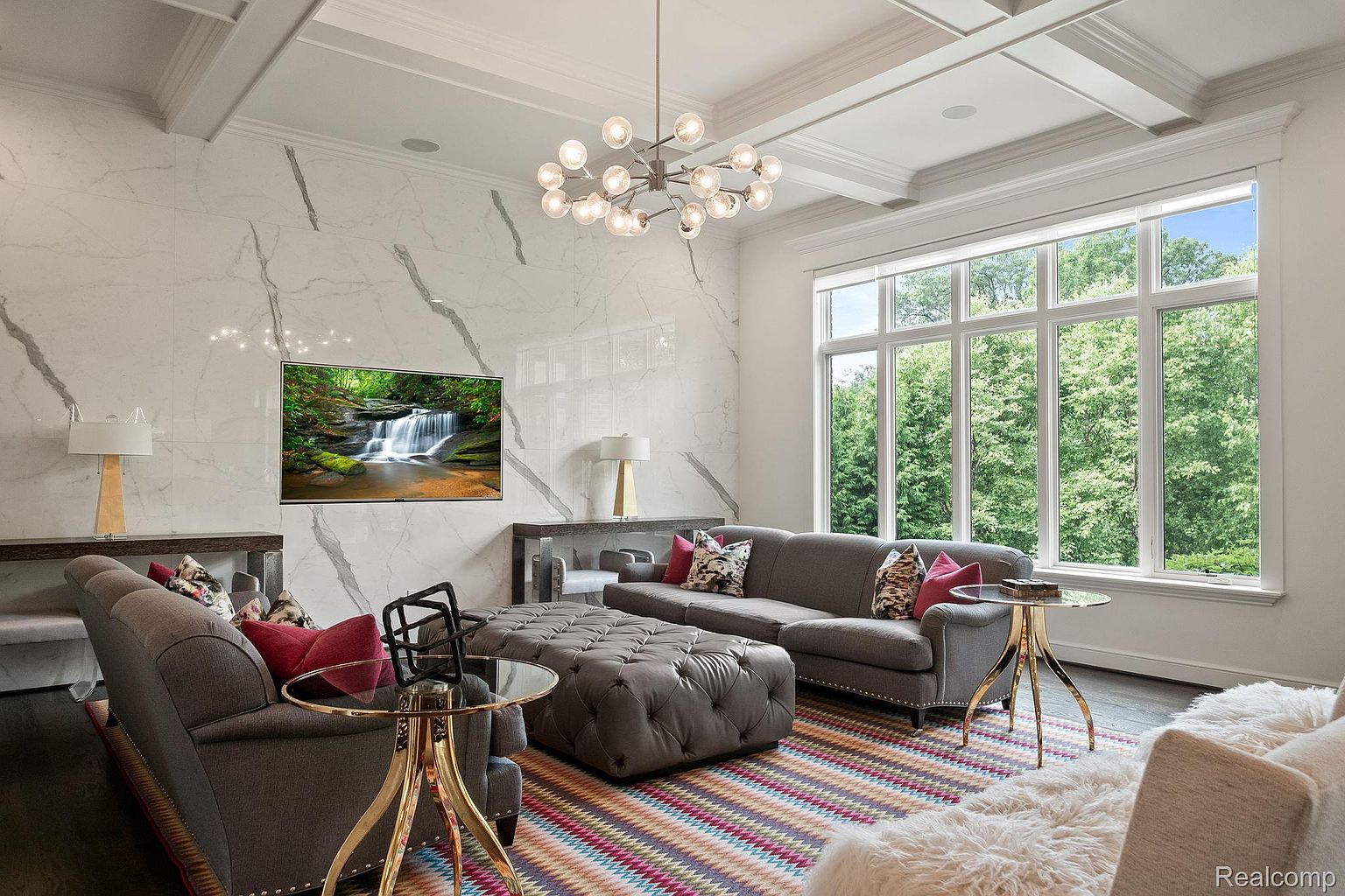 This is a well-lit living room featuring a large window with a view of lush greenery. The room is decorated with a gray sofa, a tufted ottoman, and a modern chandelier. A large television is mounted on a marble accent wall, adding a focal point to the space.