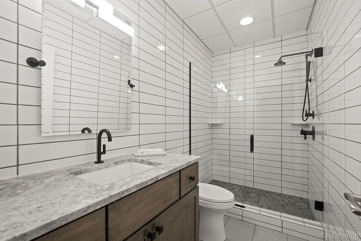 This is a modern bathroom featuring white subway tile walls with dark grout, a wood-tone vanity with a light granite countertop, and a black faucet. The shower has a glass enclosure and a black shower system. The overall design is clean and contemporary, creating a stylish and functional space.