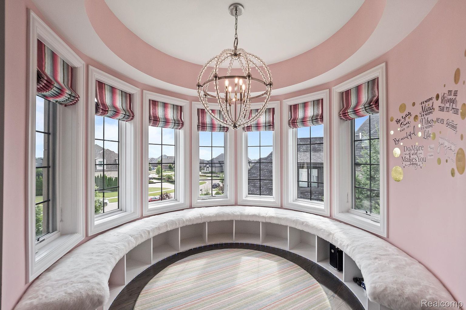 This sunroom features a curved window seat with built-in shelving, topped with a plush white cushion. The room is painted a soft pink, complemented by a striped rug and a decorative chandelier. The large windows offer ample natural light and views of the surrounding neighborhood, creating a cozy and inviting space.