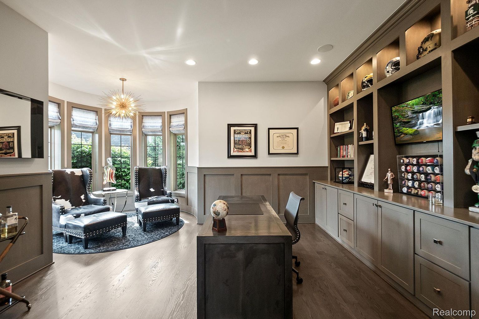This is an interior shot of a well-appointed home office. The room features a large desk, comfortable seating area with two chairs and ottomans, and custom built-in shelving with display items. The color palette is neutral with wood flooring, creating a sophisticated and inviting workspace.