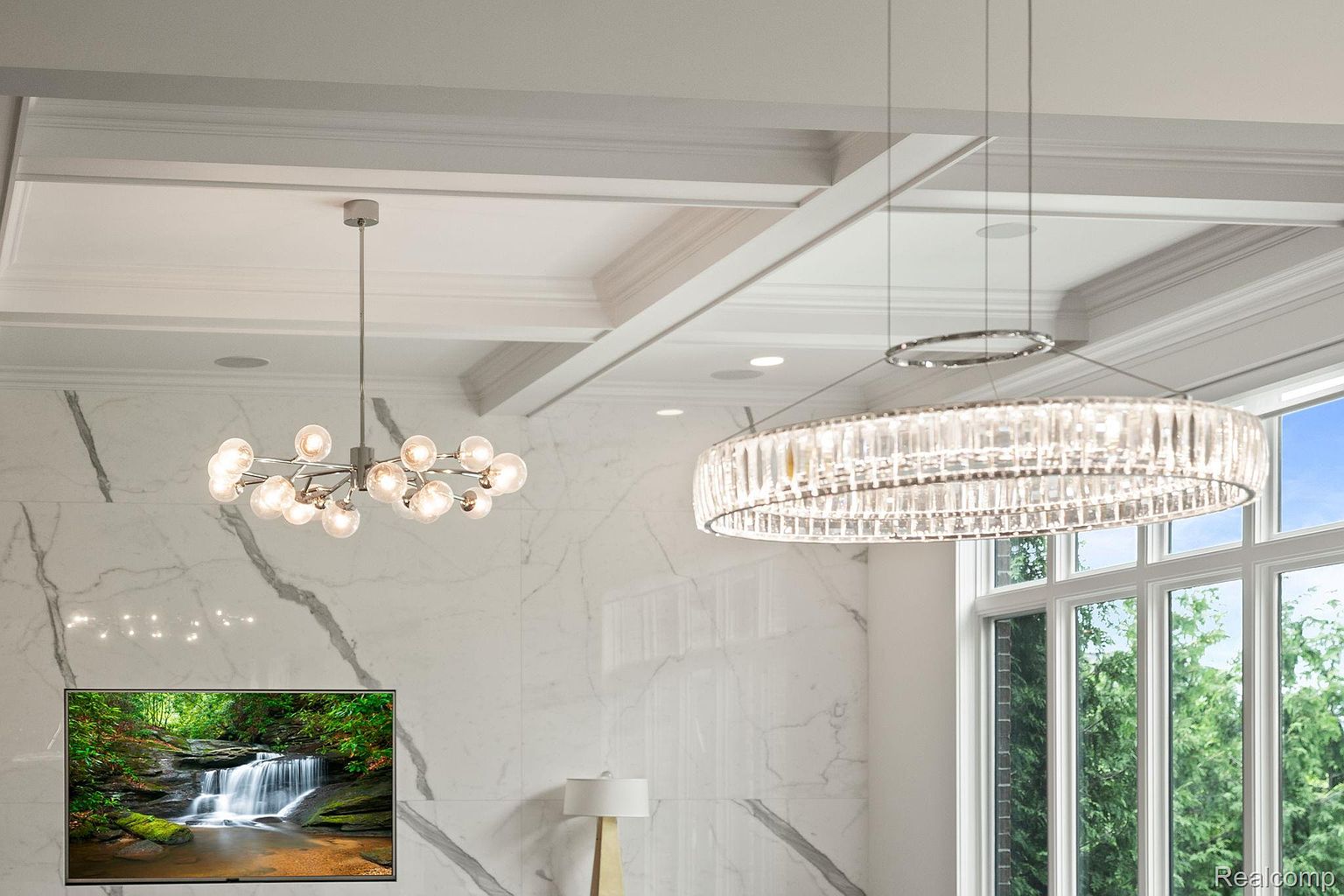 This interior shot showcases a modern living room with a focus on elegant lighting and a clean aesthetic. Two statement chandeliers hang from the coffered ceiling, illuminating the marble-clad wall and a flat-screen TV displaying a nature scene. The room exudes a luxurious and contemporary feel, perfect for a sophisticated buyer.