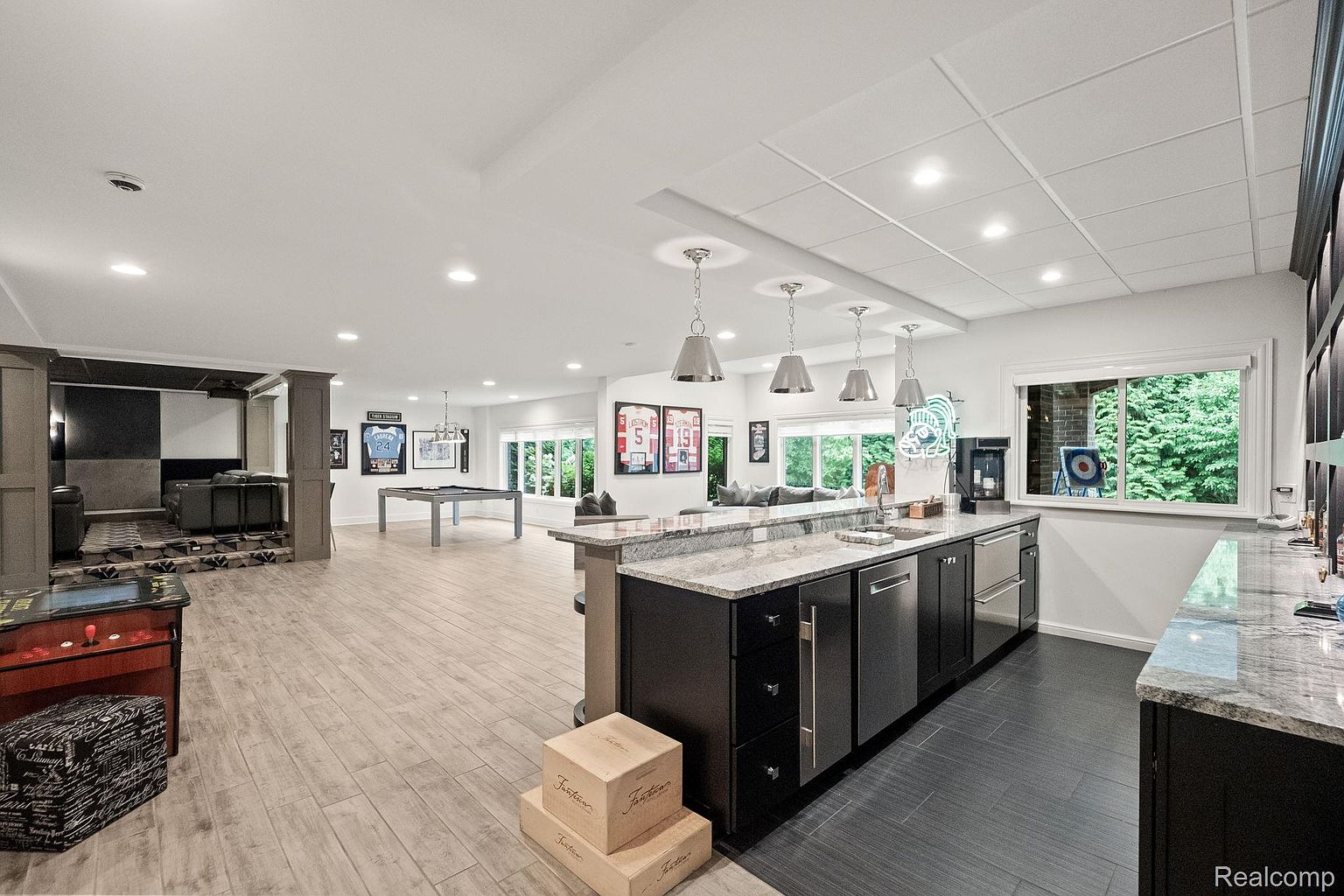 This is a spacious finished basement featuring a bar area with granite countertops, dark cabinetry, and stainless steel appliances. The basement also includes a pool table, a home theater area, and ample space for recreation and relaxation. The flooring is a light wood-look tile, and the overall design is modern and inviting.