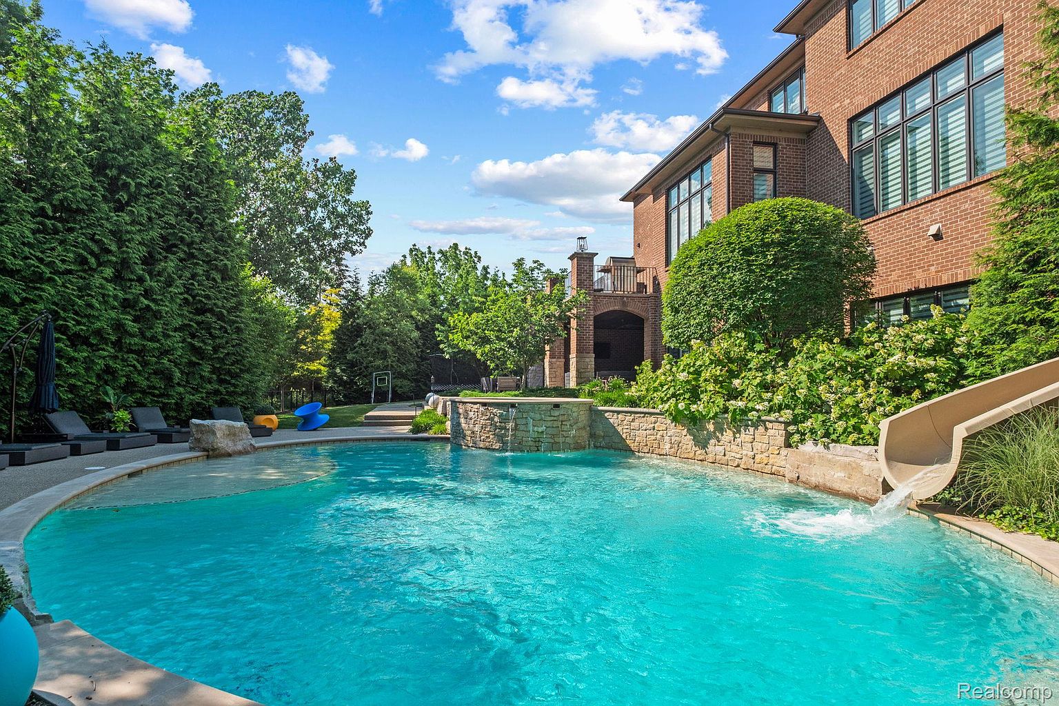 This image showcases a luxurious backyard pool area. The pool features a built-in waterfall and a slide, surrounded by lush landscaping and a stone retaining wall. A large brick house is visible in the background, suggesting a high-end property with ample outdoor entertainment space.