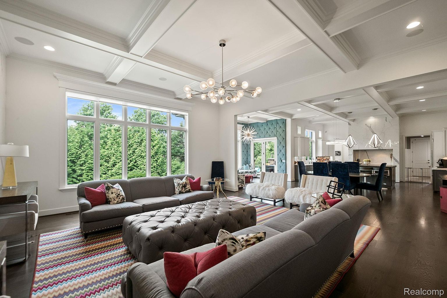 This is an interior shot of a luxurious living room featuring two gray sofas, a large tufted ottoman, and a colorful chevron-patterned rug. A large window provides ample natural light and a view of the surrounding greenery. The room is decorated with modern lighting fixtures and stylish accent pillows, creating an inviting and sophisticated atmosphere.