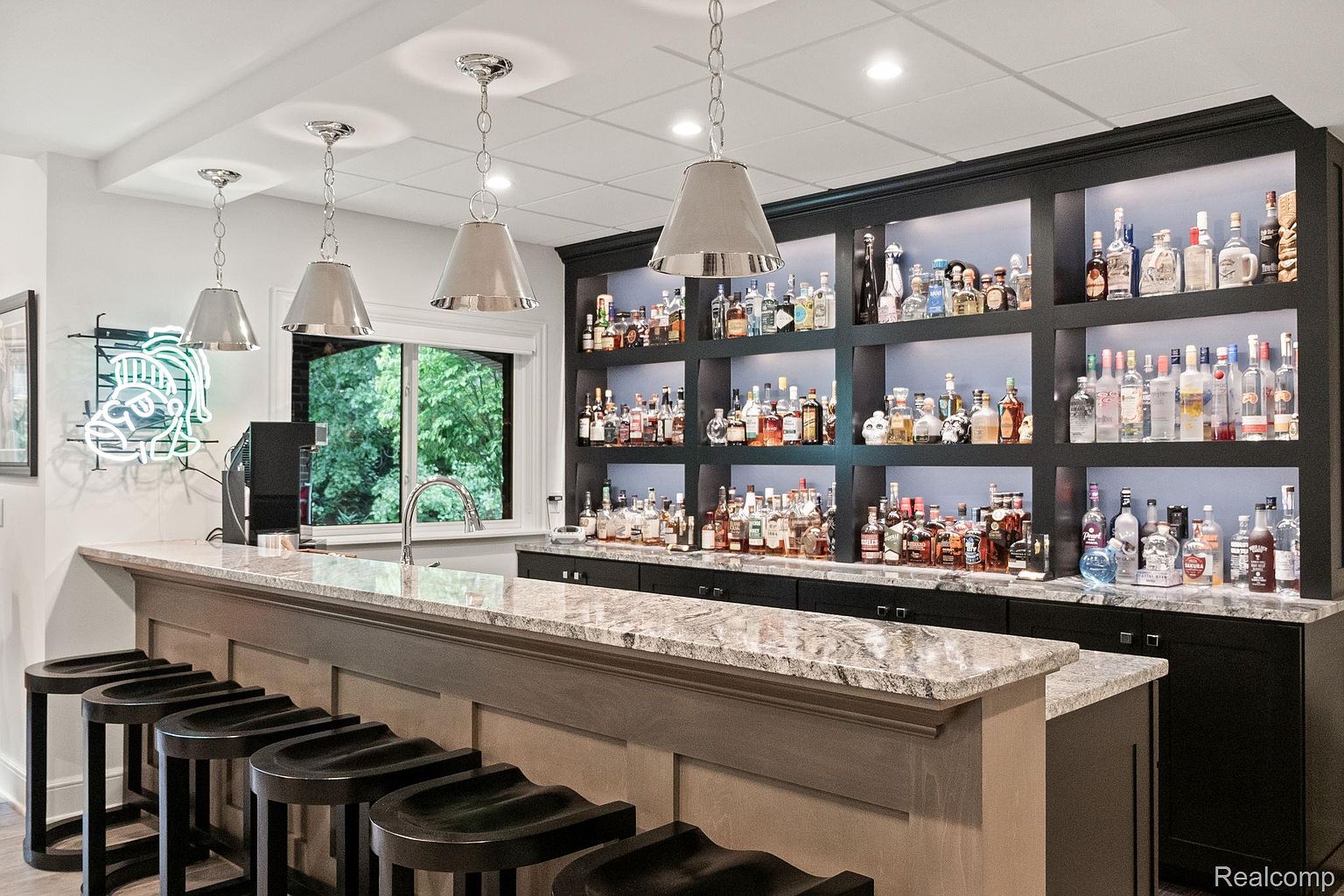 This image showcases a stylish basement bar area, complete with a granite countertop, dark wood cabinetry, and open shelving displaying an extensive liquor collection. Pendant lighting adds a touch of elegance, while bar stools provide seating for guests. The overall impression is that of a sophisticated and inviting entertainment space.