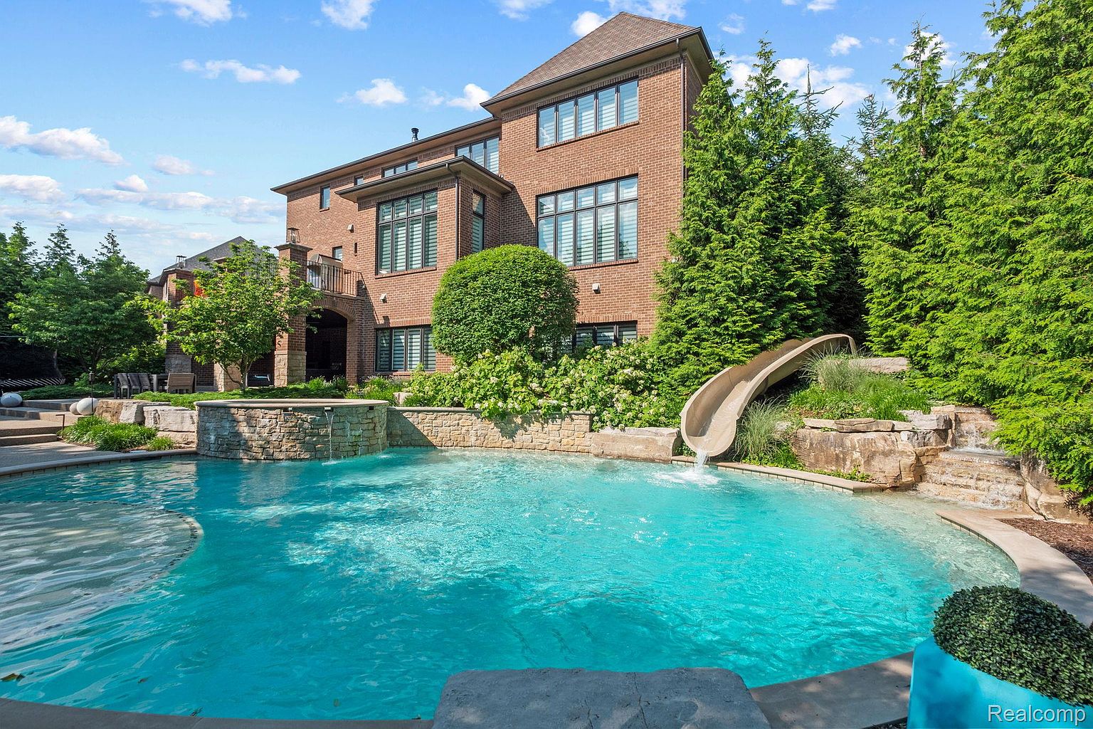 This image showcases a luxurious backyard pool area with a brick house in the background. The pool features a slide and a waterfall feature, surrounded by lush greenery and stone landscaping. The scene evokes a sense of relaxation and upscale outdoor living, perfect for entertaining or enjoying a private retreat.