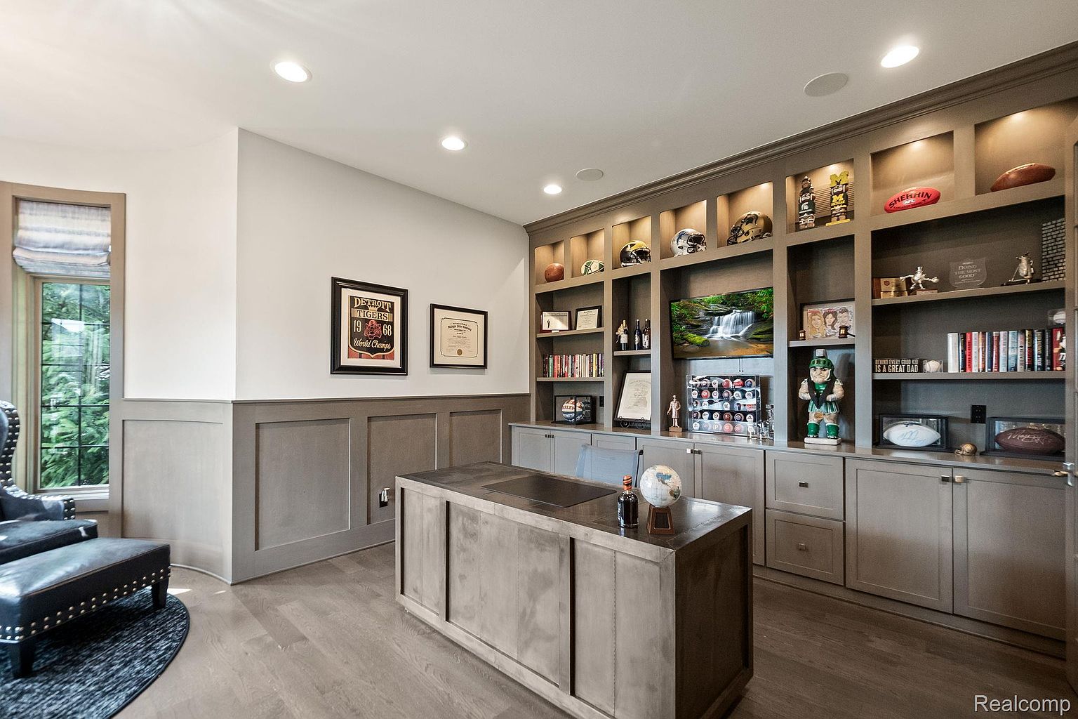 This is an interior shot of a well-appointed office or study. The room features custom built-in shelving with decorative items and a television, a large central desk, and a seating area with a leather chair and ottoman. The color palette is neutral, creating a sophisticated and professional atmosphere.