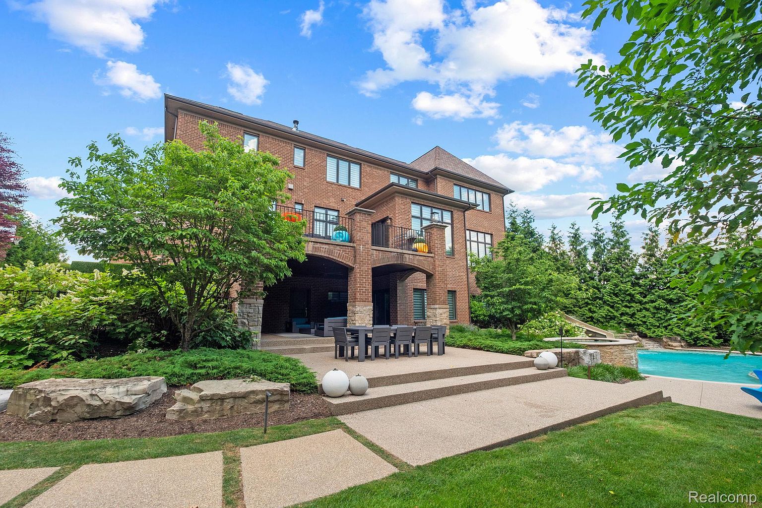 This is a rear view of a large, two-story brick house with a well-manicured backyard. The house features multiple balconies, arched entryways, and a patio with outdoor seating. A swimming pool is visible in the background, surrounded by lush greenery, creating a luxurious and private outdoor space.