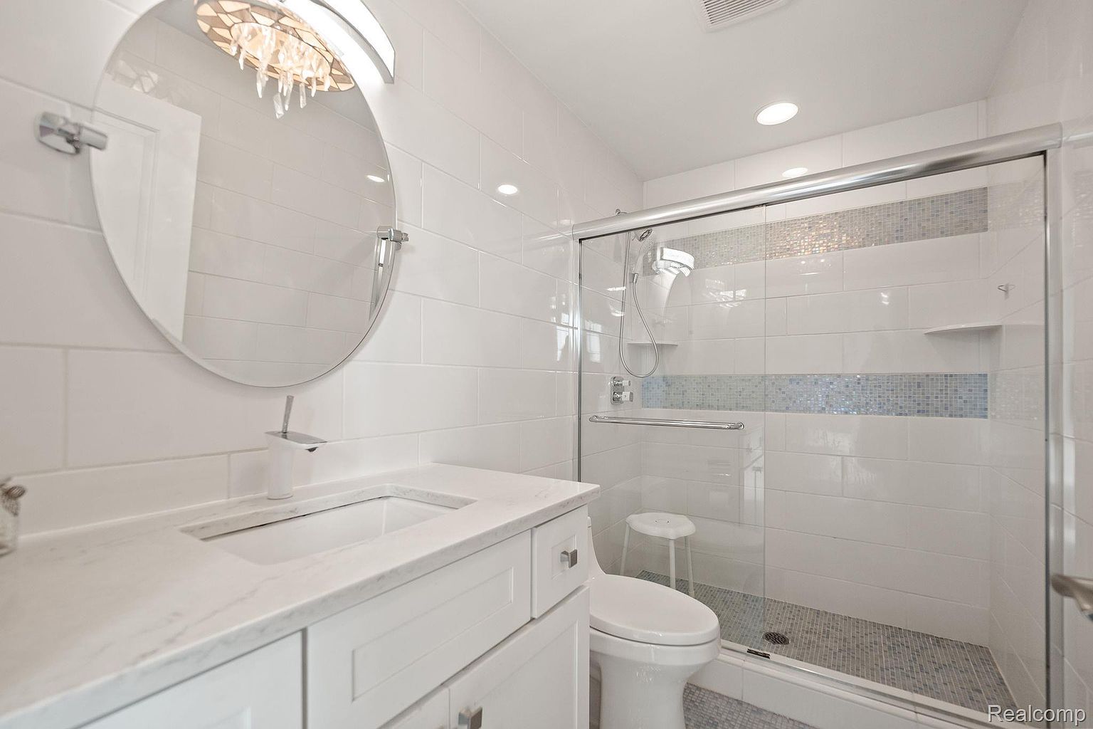 This is a bright and modern bathroom featuring white subway tiles, a round mirror with decorative lighting, and a white vanity with a marble countertop. The shower has a glass enclosure with mosaic tile accents and a built-in shelf. The overall impression is clean, stylish, and well-maintained.