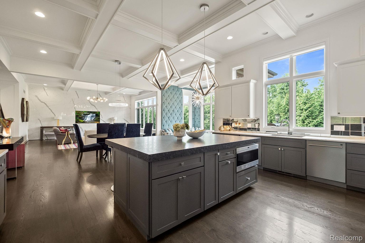 This interior shot showcases a modern kitchen with a large central island featuring dark gray cabinetry and a contrasting countertop. The kitchen seamlessly blends into an open-concept dining and living area, highlighted by elegant pendant lighting and a coffered ceiling. Large windows provide ample natural light and views of the outdoors, enhancing the spacious and luxurious feel of the home.