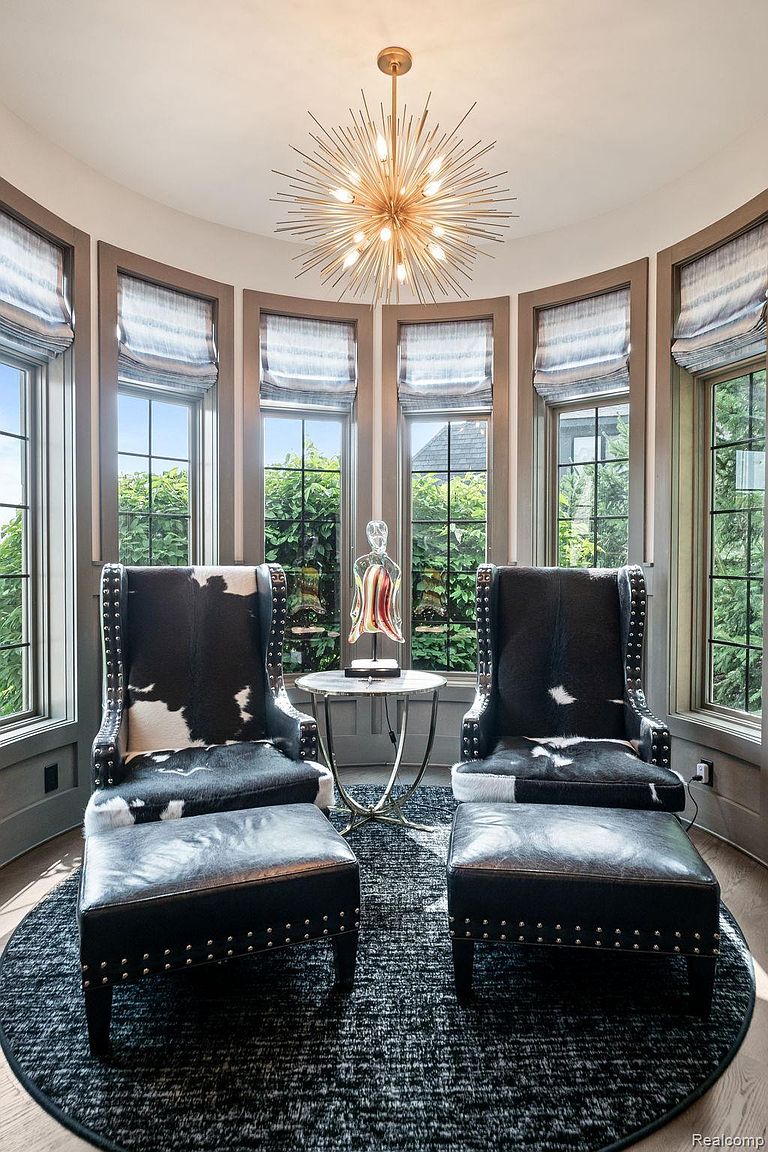 This sunroom features two black and white cowhide chairs with matching ottomans, arranged on a round patterned rug. A small round table with a decorative glass sculpture sits between the chairs. The room is surrounded by windows with blue and white striped roman shades, and a modern starburst chandelier hangs from the ceiling, creating a cozy and stylish space.
