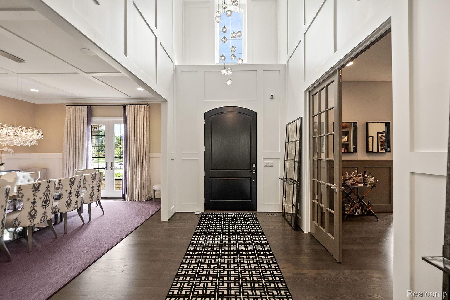 This is an interior shot of a grand hallway featuring a high ceiling with a decorative chandelier. The entryway is adorned with white wainscoting and a black front door, complemented by a geometric patterned runner rug. To the left, a dining area is visible, and to the right, an open doorway leads to another room, creating a sense of spaciousness and luxury.