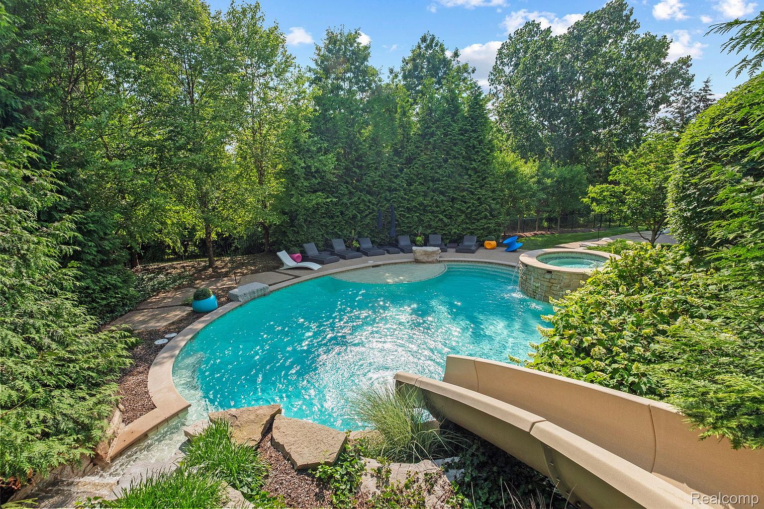 This image showcases a luxurious backyard pool and spa area. The pool features a unique curved design with a built-in spa and a water slide. Lush greenery surrounds the pool, creating a private and serene oasis, perfect for relaxation and entertainment.