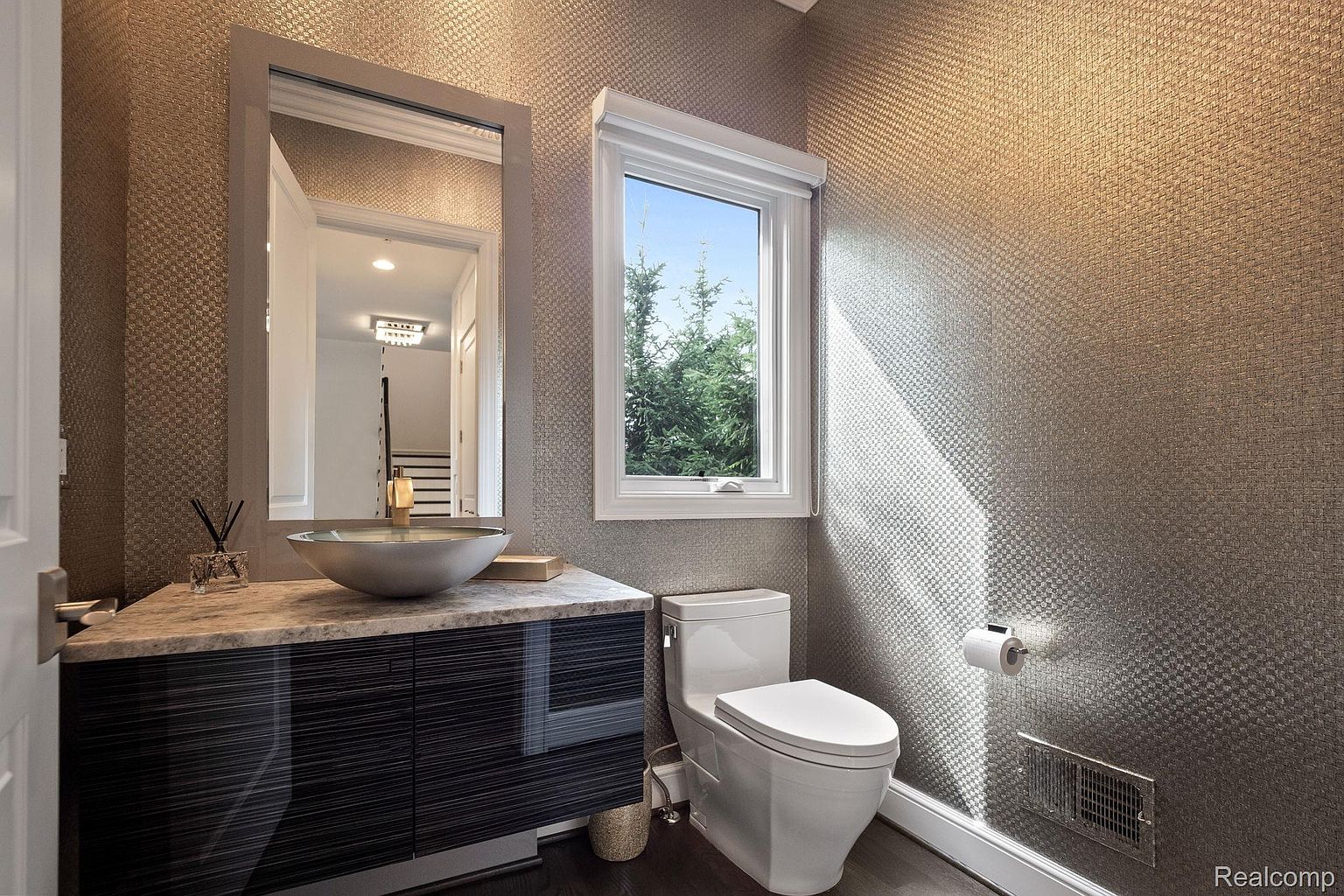 This is a well-lit guest bathroom featuring textured wallpaper, a modern vessel sink on a dark wood vanity with a marble countertop, and a toilet. A window provides natural light and a view of greenery outside. The overall impression is clean, stylish, and functional.