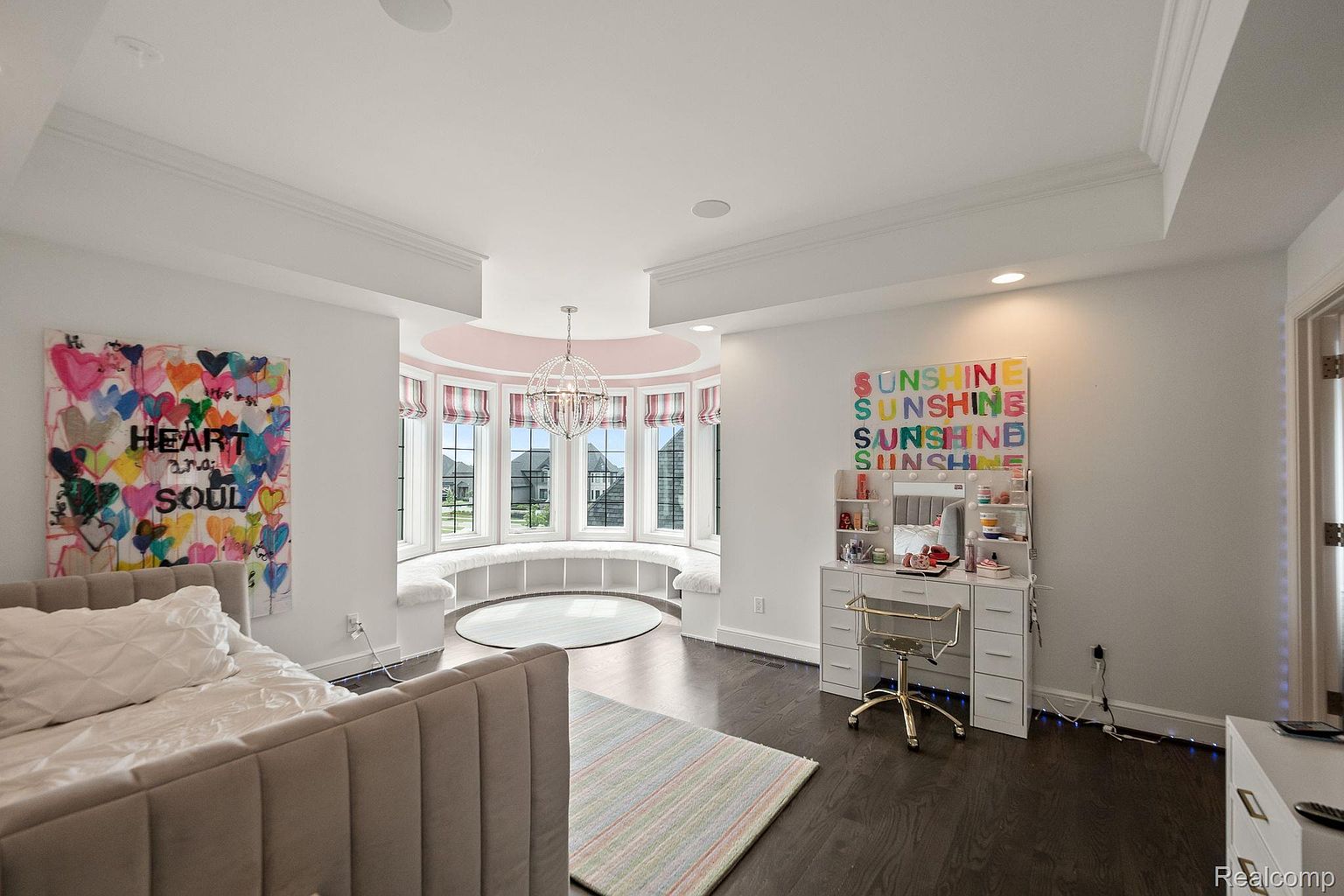 This is a bright and stylish bedroom featuring a large bay window with a built-in seating area, a modern chandelier, and a playful art piece on the wall. The room includes a vanity area with a gold chair and a bed with a tufted headboard. The dark hardwood floors contrast nicely with the white walls and trim, creating a clean and inviting space.