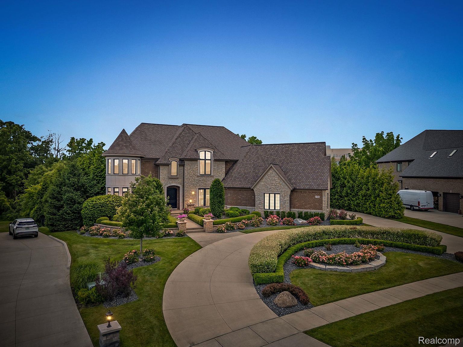 This is a stunning front view of a large, luxurious home. The house features a combination of stone and brick exterior, a turret-style architectural element, and a well-manicured lawn with mature trees and flower beds. A circular driveway leads to the entrance, enhancing the property's curb appeal and creating a grand impression.